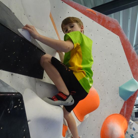 Ein junger Junge klettert an einer Wand in einem Indoor-Kletterzentrum. Er trägt ein grün-gelbes Shirt und schwarze Shorts.