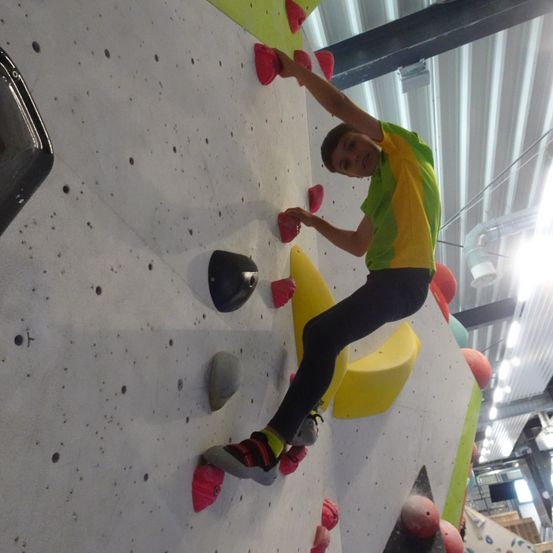 Eine Person klettert an einer Wand mit bunten Griffen. Sie trägt ein grün-gelbes Shirt und schwarze Hose. Die Wand ist weiß mit verschiedenen farbigen Griffen.
