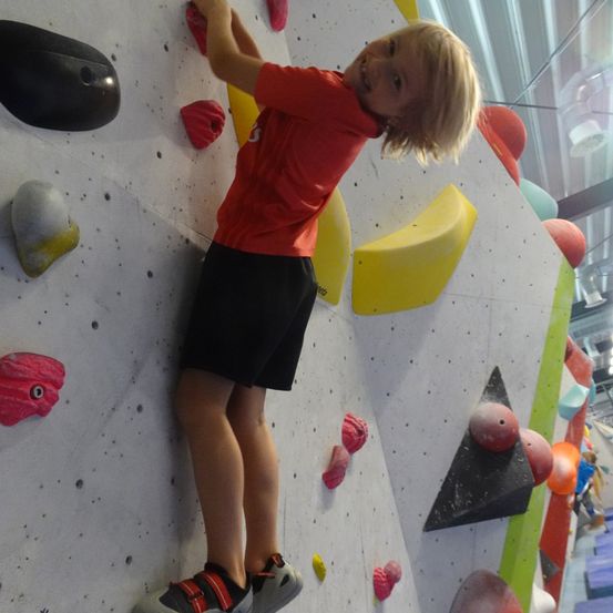 Ein Kind in einem roten T-Shirt und schwarzen Shorts klettert an einer Indoor-Kletterwand mit verschiedenen farbigen Griffen. Die Wand ist mit unterschiedlichen Formen und Farben bedeckt.
