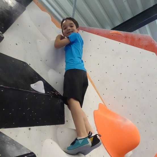 Ein junger Junge klettert an einer Wand mit künstlichen Griffen hoch. Er trägt ein blaues Shirt, schwarze Shorts und Turnschuhe.
