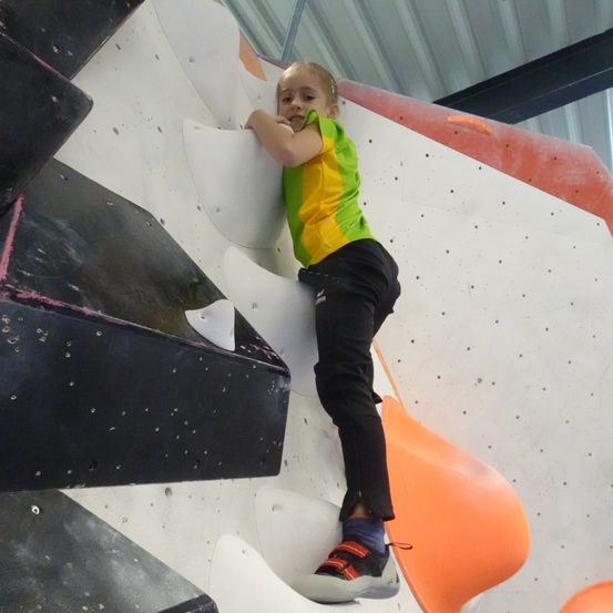 Ein junges Mädchen klettert an einer Kletterwand, trägt ein grün-gelbes Shirt, schwarze Hose und Turnschuhe. Die Wand hat verschiedene farbige Griffe.