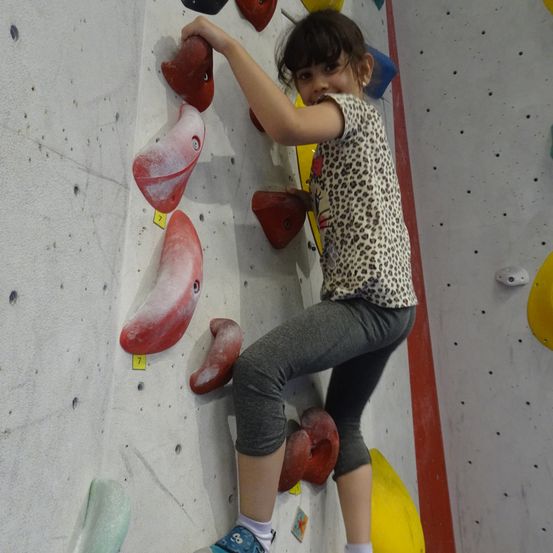 Ein kleines Mädchen klettert an einer Wand mit ihren Händen an roten Griffen, trägt ein Leopardenmuster-Shirt und graue Hose. Die Wand hat verschiedene farbige Griffe und es gibt kleine gelbe Aufkleber mit Nummern.