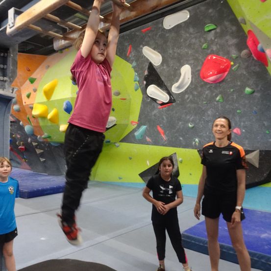 Ein junges Mädchen hängt in einem Fitnessstudio von einem Klettergriff an einer farbenfrohen Kletterwand, während zwei Frauen zusehen.