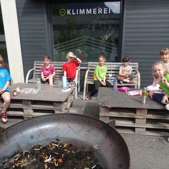 Mehrere Kinder sitzen auf Bänken vor einem Gebäude mit dem Schild 'Klimmerei'. Sie haben Gegenstände auf einem Tisch und eine große Schüssel mit ausgelöschten Zigaretten.