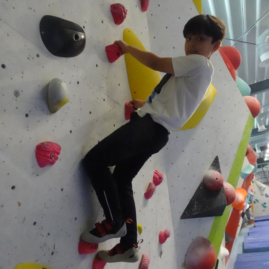 Ein junger Junge klettert an einer Wand mit verschiedenen farbigen Griffen und Haltepunkten und trägt ein weißes T-Shirt und schwarze Hose.