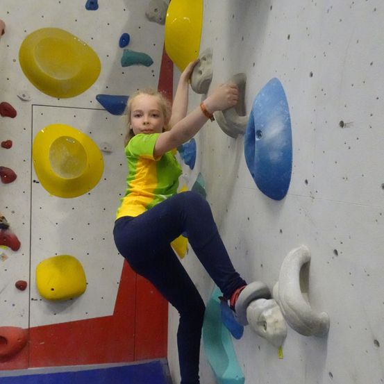 Ein kleines Mädchen in einem grünen Shirt und blauen Hosen klettert an einer Wand mit verschiedenen farbigen Griffen.