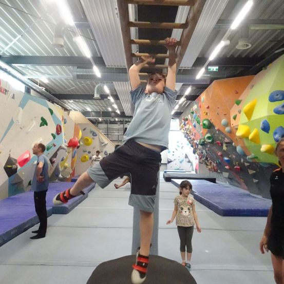 Ein Junge klettert eine Holzleiter in einem Fitnessstudio mit Kletterwänden, andere beobachten.