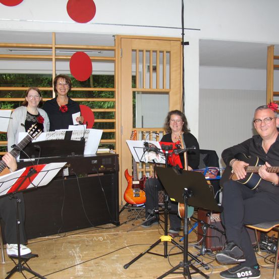 Eine Gruppe von Menschen spielt Gitarren und singt in einem Raum mit einer Holzwand. Ein Mann sitzt und lächelt, während er eine Gitarre spielt. Die anderen stehen und schauen auf ihre Notenblätter.