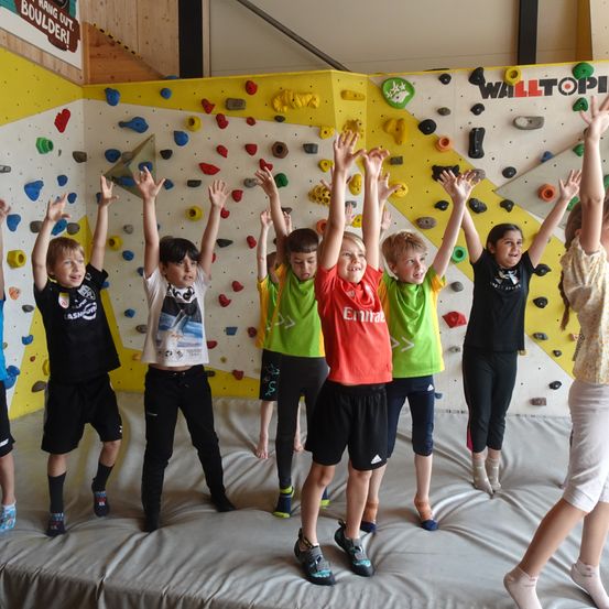 Kinder in verschiedenen farbigen Shirts und Hosen stehen auf einer Matte und heben ihre Hände, möglicherweise in einem Kletterzentrum.