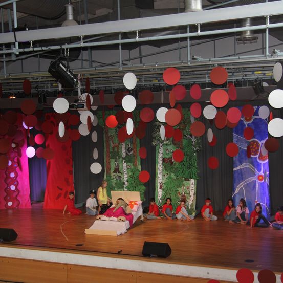 Kinder führen ein Theaterstück auf einer Bühne mit buntem Hintergrund, kreisförmigen Dekorationen und einem Bett auf. Ein Kind liegt auf dem Bett, während andere auf dem Boden sitzen.