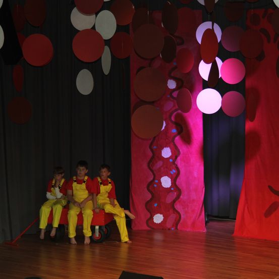 Drei Kinder in gelben Overalls sitzen auf einem roten Wagen auf einer Bühne mit rotem Hintergrund und farbenfrohen kreisförmigen Dekorationen.