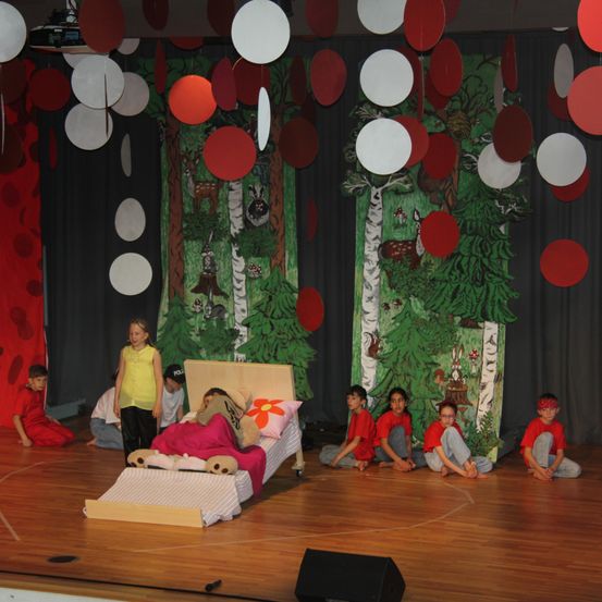 Kinder führen ein Theaterstück auf einer Bühne mit einem Bett, roten und weißen Luftballons und einem Waldhintergrund auf. Ein Kind liegt auf dem Bett, während andere herumsitzen.