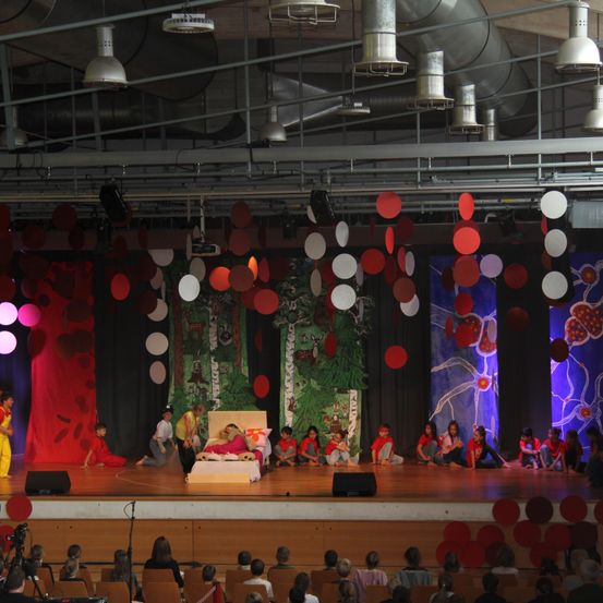 Eine Bühnenaufführung mit Schauspielern in roten Kostümen, die auf einem Bett und darum herum sitzen, rote und weiße Luftballons hängen von der Decke, und ein Publikum schaut zu.