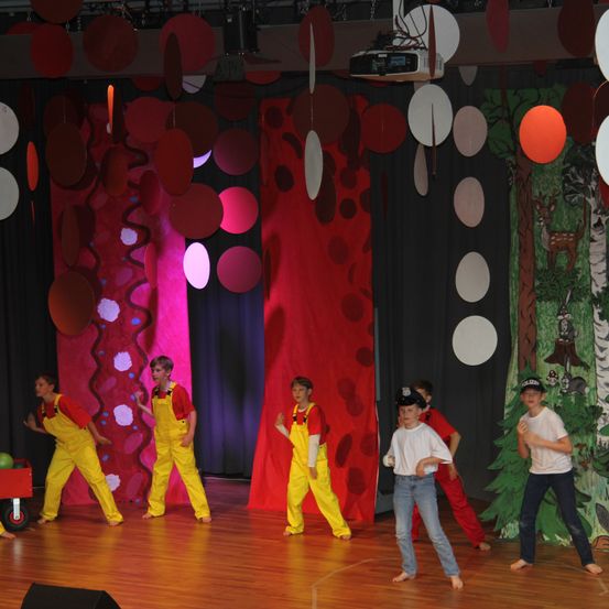 Eine Gruppe von Kindern tritt auf einer Bühne auf. Einige sind in gelben Overalls gekleidet, während andere weiße Hemden und blaue Jeans tragen. Ein farbenfroher Hintergrund mit verschiedenen Formen und Farben steht hinter ihnen.