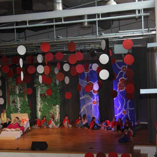 Eine Bühne mit einem Bett und roten und weißen Kreisen, die darüber hängen, mit Kindern, die vor einer Kulisse mit Bäumen und einem Tier auf dem Boden sitzen.