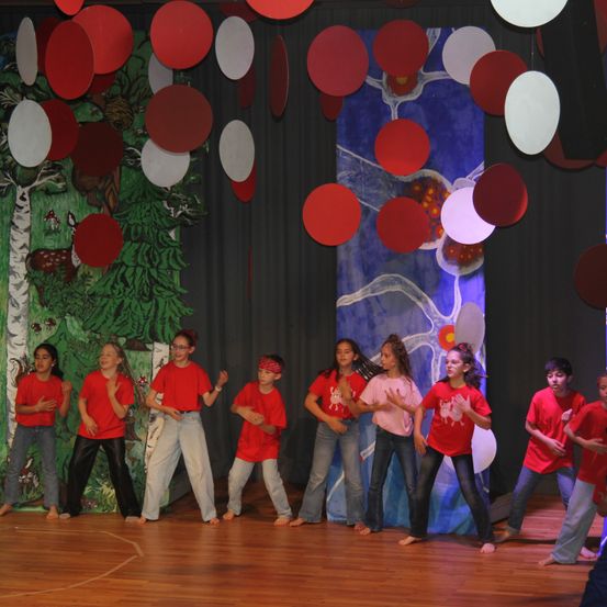Eine Gruppe von Kindern in roten T-Shirts und Jeans führt einen Tanz auf einer Bühne mit einer Kulisse aus Bäumen und Ballonen auf.