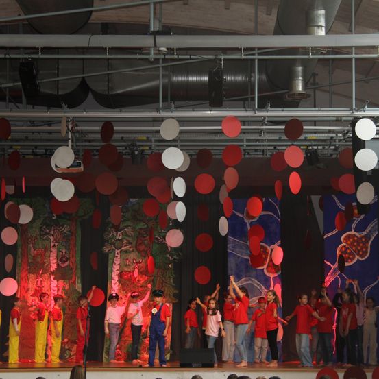 Eine Gruppe von Kindern in roten Kostümen tritt auf der Bühne auf, über ihnen schweben rote und weiße Luftballons.