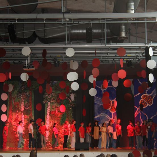 Eine Gruppe von Kindern in roten und weißen Kostümen tritt auf einer Bühne auf, an der rote und weiße Luftballons hängen. Der Bühnenhintergrund zeigt Bäume und Wildtiere.