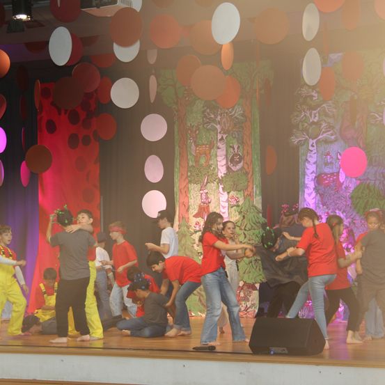 Eine Gruppe von Kindern tritt auf der Bühne auf, einige in roten T-Shirts, andere in Kostümen. Die Bühne ist mit bunten runden Dekorationen und Hintergründen mit Bäumen und Tieren geschmückt.