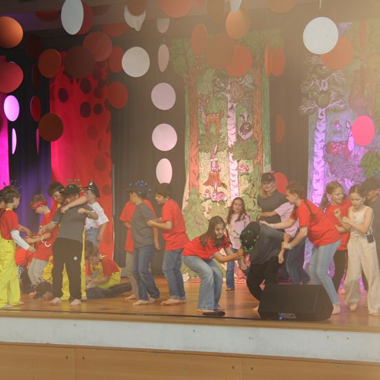 Eine Gruppe von Kindern in roten Shirts und gelben Hosen tritt auf einer Bühne mit bunten Luftballons und Kulissen auf. Ein Lautsprecher ist auf der Bühne.