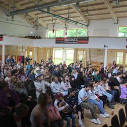 Eine große Indoor-Versammlung von Kindern und Erwachsenen, möglicherweise in einer Werkstatt, mit einem Banner, auf dem 'Bickel' und 'Spenglerrei' steht. Der Raum hat Holzwände und eine hohe Decke mit hängenden Lampen.