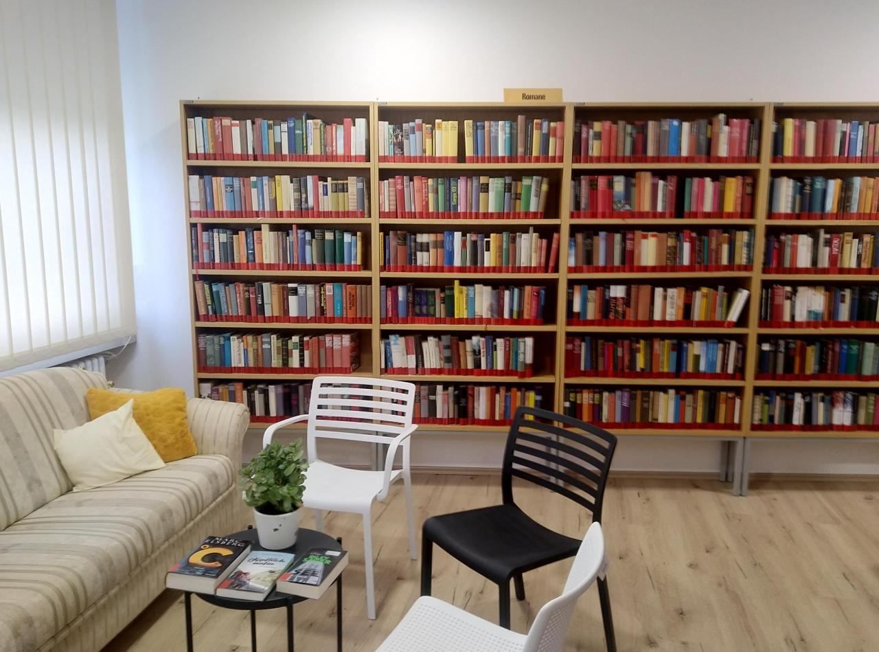 Ein Lesebereich mit einer Couch, zwei weißen Stühlen und einem schwarzen Stuhl. Ein Tisch hat drei Bücher. Ein Bücherregal ist mit Büchern gefüllt und hat ein Schild mit der Aufschrift 'Romane'.