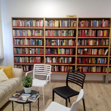 Ein Lesebereich mit einer Couch, zwei weißen Stühlen und einem schwarzen Stuhl. Ein Tisch hat drei Bücher. Ein Bücherregal ist mit Büchern gefüllt und hat ein Schild mit der Aufschrift 'Romane'.