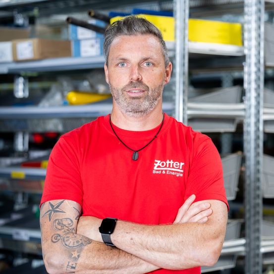 Ein Mann mit Tätowierungen an den Armen, der ein rotes Shirt mit dem Wort Zotter trägt, steht mit verschränkten Armen in einem Lagerhaus.