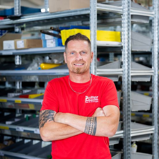 Ein Mann in einem roten Shirt mit dem Wort Zotter steht mit verschränkten Armen in einem Lagerhaus.