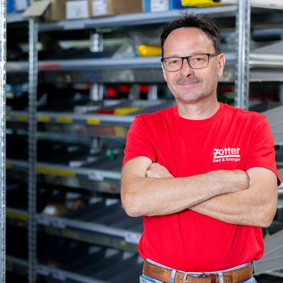 Ein Mann in einem roten Shirt mit Zotter Bad & Energie Logo steht in einem Lagerhaus mit verschränkten Armen.