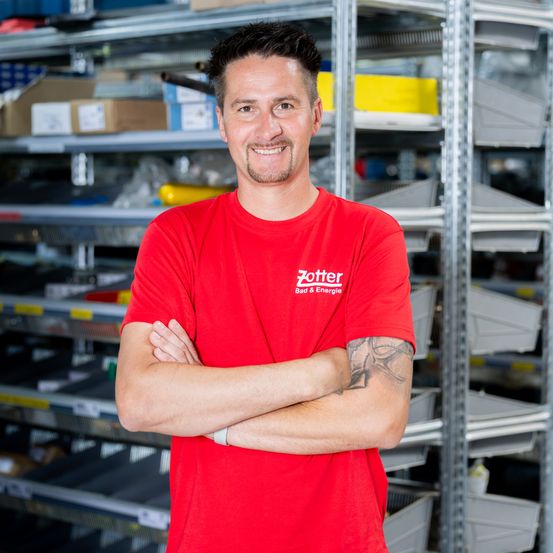 Ein Mann in einem roten Shirt mit dem Wort Zottel steht mit verschränkten Armen in einem Lager.