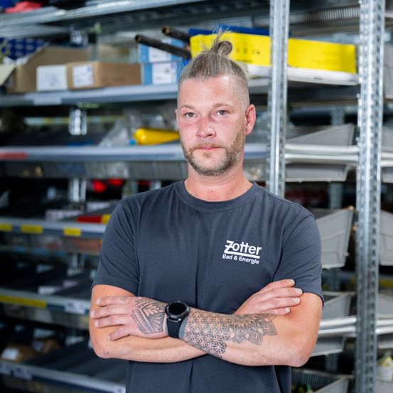 Ein Mann mit Duttfrisur und Tätowierungen an den Armen steht mit verschränkten Armen in einem Lager. Er trägt ein graues T-Shirt mit dem Text 'Zotter Bad & Energie'. Hinter ihm sind Regale mit verschiedenen Gegenständen zu sehen.