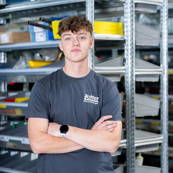 Ein junger Mann mit braunen Haaren steht in einem Lagerhaus, trägt ein graues T-Shirt mit dem Text 'Zotter Bau & Energie' und eine Smartwatch. Er hat die Arme verschränkt und schaut direkt in die Kamera. Hinter ihm befinden sich Regale mit verschiedenen Artikeln.