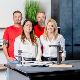 Vier Personen stehen lächelnd vor einer Arbeitsplatte für ein Foto. Zwei Frauen tragen weiße Blusen und zwei Männer rote Hemden. Eine Pflanze und ein Wasserhahn sind auf der Arbeitsplatte.