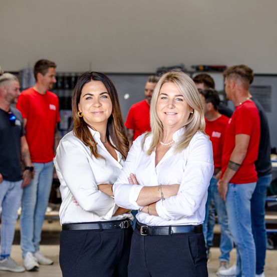 Zwei Frauen in weißen Blusen und schwarzen Hosen stehen nebeneinander mit verschränkten Armen und lächeln in einem Raum mit anderen Personen in roten Hemden.