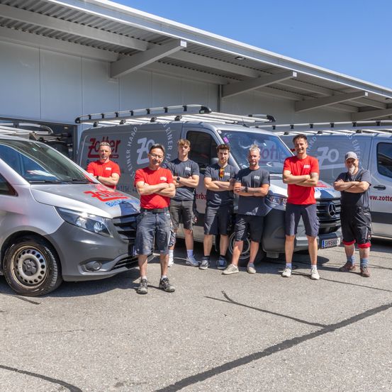 Eine Gruppe von Männern in roten und grauen Shirts steht vor drei mit Logos versehenen Lieferwagen, möglicherweise ein Teamfoto.
