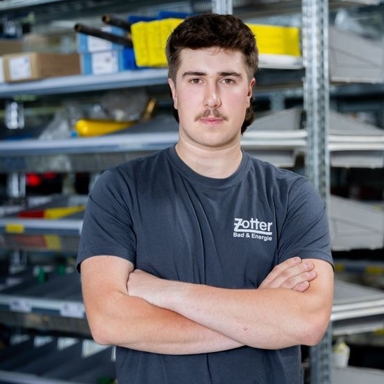 Ein Mann mit einem Schnurrbart steht in einem Lagerhaus, trägt ein graues T-Shirt mit dem Text 'Zotter Bad & Energie'. Er hat die Arme verschränkt und schaut direkt in die Kamera. Hinter ihm befinden sich Regale mit verschiedenen Gegenständen.
