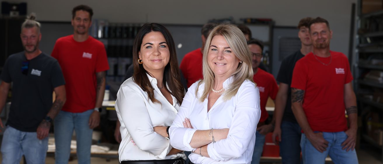 Zwei Frauen in weißen Blusen stehen selbstbewusst und lächeln für ein Foto in einer industriellen Umgebung, mit Kollegen in roten Hemden im Hintergrund.