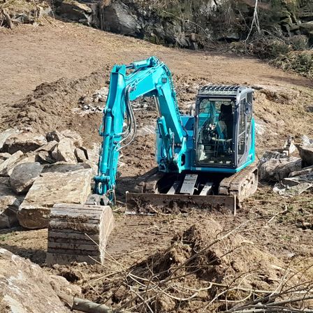 Ein blauer Bagger arbeitet auf einer Baustelle mit Erdboden, Steinen und Schutt unter einem bewölkten Himmel.