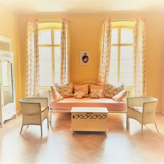 Ein gemütliches Wohnzimmer mit gelber Wand, einem Tagesbett mit rosa Laken und zwei Korbstühlen. Der Raum hat einen Holzboden und zwei Fenster mit Blumenvorhängen.