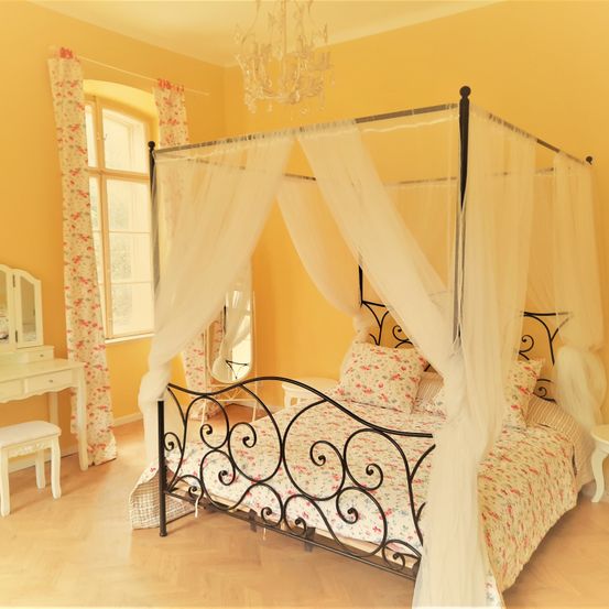 Ein Schlafzimmer mit einem Himmelbett, gelben Wänden und Blumenvorhängen. Eine Kristallleuchte und ein Spiegel sind sichtbar.