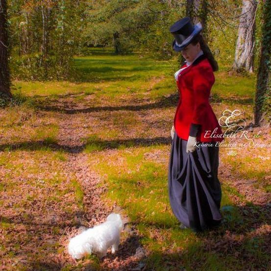 Eine Frau in einem roten Mantel und schwarzem Hut steht in einem Wald mit einem kleinen weißen Hund an ihrer Seite.