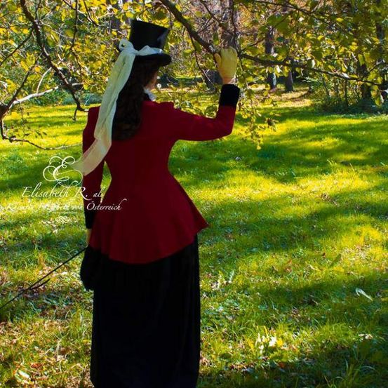 Eine Frau in einem roten Mantel und schwarzem Hut mit Handschuhen pflückt Früchte von einem Baum in einem Grasfeld.