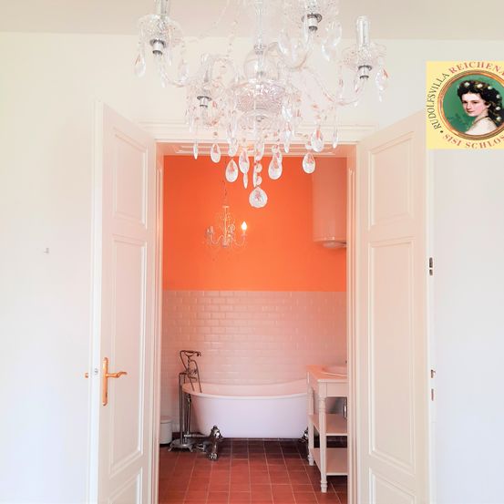 Ein Badezimmer mit einer weißen Badewanne, einer orangefarbenen Akzentwand und einer Kristallleuchte, die von der Decke hängt.
