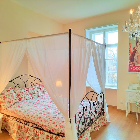 Ein Schlafzimmer mit einem Baldachinbett, das mit Blumenbettwäsche verziert ist, einer Kristallleuchte und einem Fenster mit Blick auf Bäume.