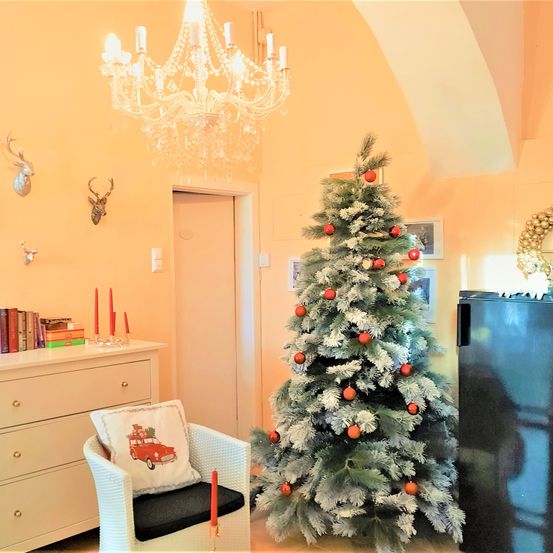 Ein gemütliches Zimmer mit einem Weihnachtsbaum, einem weißen Stuhl mit Kissen und einer Kristallleuchte. Ein weißer Schrank mit Büchern und Kerzen steht links.