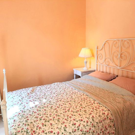 Ein Schlafzimmer mit einem weißen Metallbettgestell, einem Blumenbettüberwurf und pfirsichfarbenen Wänden. Eine weiße Lampe steht auf dem Nachttisch.