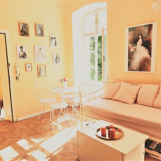 Ein gemütliches Wohnzimmer mit einem Sofa, einem kleinen Tisch und einem Fenster mit Blick auf Bäume. Die Wände sind gelb gestrichen und es hängen mehrere gerahmte Bilder an der Wand.