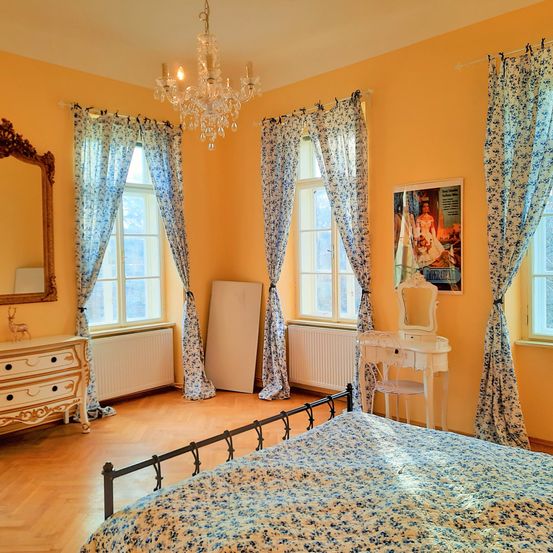 Ein Schlafzimmer mit einem Holzbett, Blumenbettwäsche, Kronleuchter und gelben Wänden. Ein Spiegel, eine Kommode und ein Schreibtisch sind ebenfalls vorhanden.