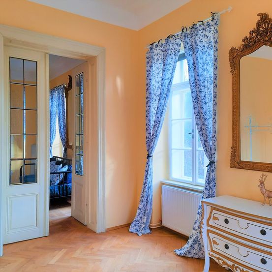Ein elegantes Zimmer mit Holzboden, blauen Vorhängen und einer weißen Kommode mit einer Hirschfigur. Der Raum hat einen Spiegel und ein Fenster mit einem Heizkörper darunter.